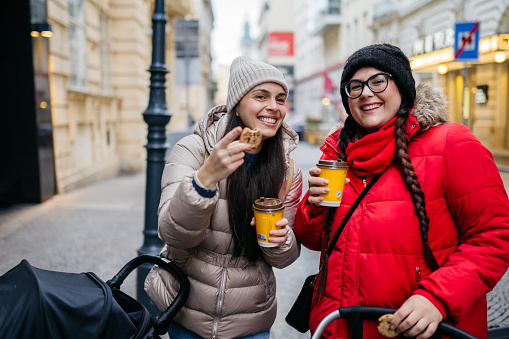 Two mothers are walking together on the sidewalk with a cup of coffee