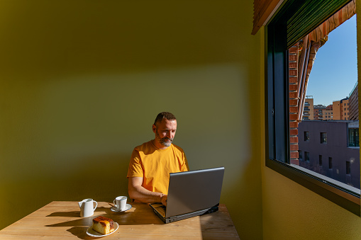 Alcorcón, Madrid, Spain. Smiling mature man working from home with his laptop by the sunny window.