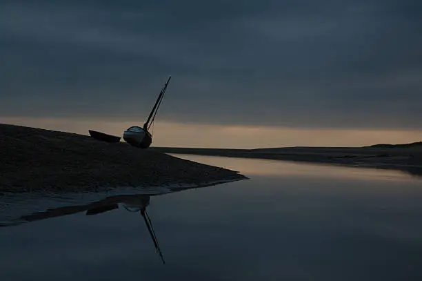 Photograph of a sailing boat beached at low tide on the Norfolk coast. Photo was taken shortly after dawn on a cloudy day in summer.