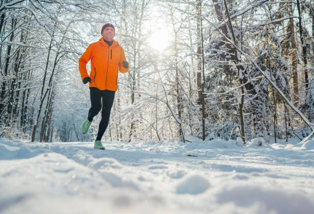 istock Middle-aged smiling trail runner man dressed bright orange windproof jacket endurance fast running picturesque snowy forest during sunny frosty day. Sporty active people and winter training image. 1855977764