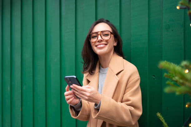 Close up smiling woman using smartphone, standing near Christmas tree