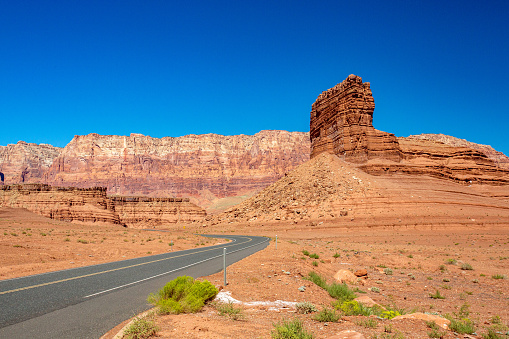 Highway near Marble Canyon in the Arizona Desert. The road winds around a mountain range with unique geological features. The stone face is red and is in contrast with the deep blue cloudless sky. There are small green shrubs in the foreground, with stones that are red and orange, with a tall monolithic structure. There is plenty of copy space in the sky above, a perfect tourist vacation landscape of this rural beautiful nature scene.
