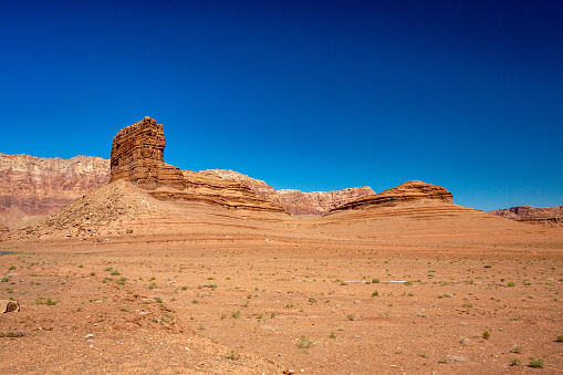Remote and Barren Desert Landscape in Arid Arizona, the red and orange desert landscape has small green brush plants in the foreground, with a rough and eroded cliff face in the sandstone of this scenic valley. The desolation and barrenness make it look like an alien landscape with a deep blue cloudless sky in the background. A great tourism photo for the southeast.