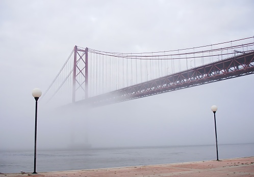 The Lisbon landmark, 25 de Abril Bridge, veiled in the mist rising from the Tagus River at dawn, creating an ethereal and captivating ambiance that adds a touch of mystique to the cityscape.