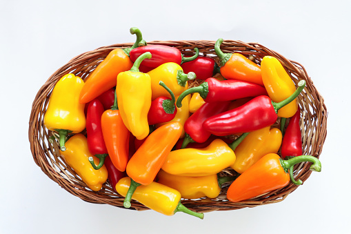 Stock photo showing close-up, elevated view of white background with a heap of red, orange and yellow mini peppers (Capsicum annuum), with green stems in a wicker basket.