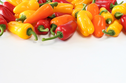 Stock photo showing close-up view of white background with a small heap of red, orange and yellow mini peppers (Capsicum annuum), with green stems.
