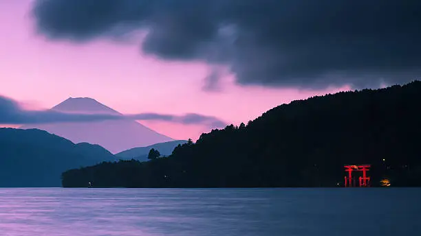 A torii gate is lit up on the shoreline, while a pink and purple sun sets over Mount Fuji in the backdrop.