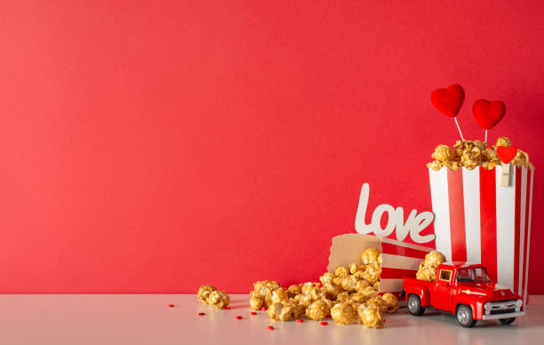 Valentine's Day special premiere for hopeless romantics. Side view table set with tiny car, popcorn spilled from striped boxes, heart decor, sprinkles. Red wall backdrop creates enchanting ambiance