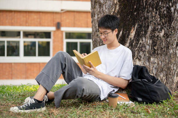 un estudiante asiático nerd y positivo está escribiendo algunas ideas en un libro debajo del árbol en un parque del campus. - male education college student nerd fotografías e imágenes de stock
