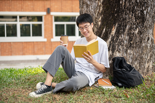 A positive young Asian male college student is sipping coffee and reading a book under the tree in a campus park. Lifestyle, Uni life