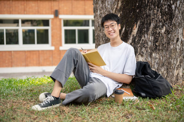 un estudiante asiático nerd y positivo está escribiendo algunas ideas en un libro debajo del árbol en un parque del campus. - male education college student nerd fotografías e imágenes de stock
