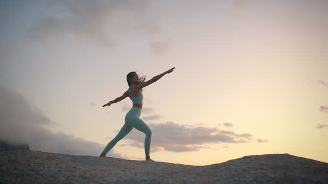 Woman, yoga and warrior pose on beach at sunset for mindfulness practice, balance lotus or healthy self care. Female person, rock and spiritual zen in nature or peace, wellness or healing aura
