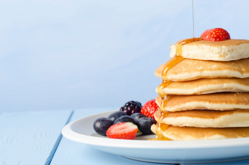 A stack of breakfast pancakes topped with a strawberry, standing on a white china plate, surrounded by Summer fruits, with maple syrup being poured from above and drizzling over the pancakes.  Pale blue wooden table and background.