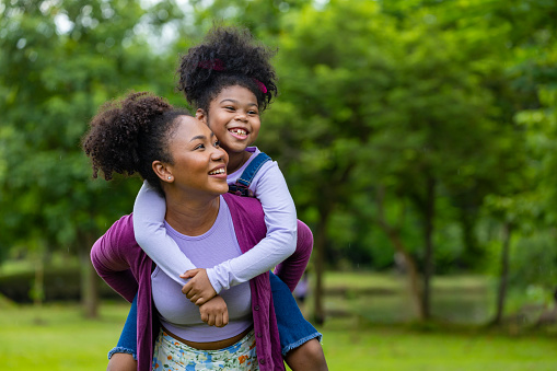 African American mother is playing piggyback riding with her young daughter while having a summer picnic in public park for wellbeing and happiness