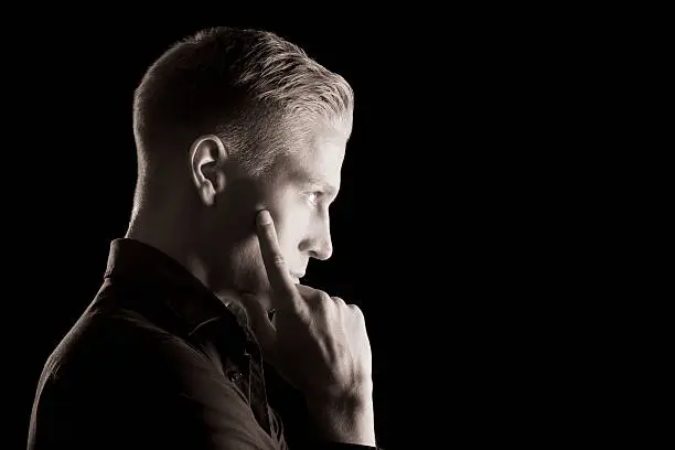 Side-face low key portrait of young attractive man in dark shirt with hand at chin looking straight, black and white, isolated on black background.