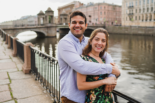 Embraced couple smiling by the river, an iconic bridge in the backdrop. Young man with wife at travel, vacation trip, honeymoon.