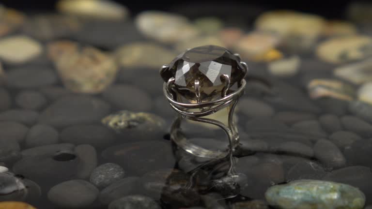 Drops of water fall on the silver ring with smoky quartz, that is in water with black pebbles, slow motion