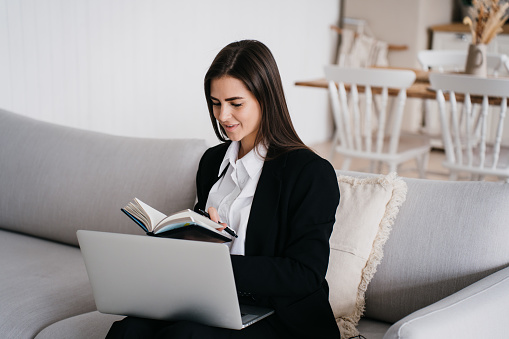 diligent young woman in professional attire is engrossed in her research on a laptop, with a notebook in hand, seated on a chic sofa in a well-appointed, airy room.