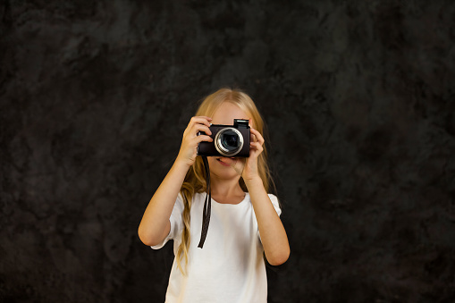 Portrait of girl child model in white t-shirt a taking photo, posing at camera. Positive cover kid 6 year old with photo camera at dark black, studio shot. Kids emotional concept. Copy ad text space