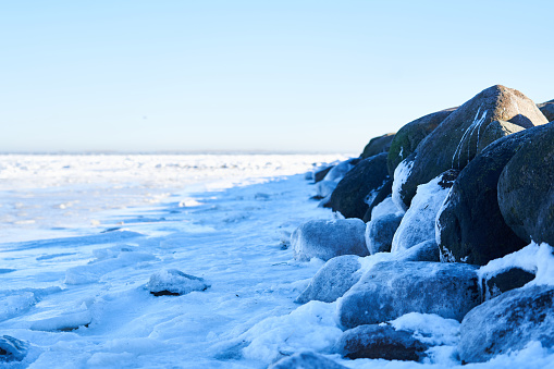 Rocks stacked by snow covered shore at beach against sky on sunny day in winter