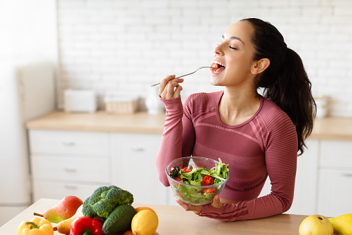 Tasty Weight Loss. Fit lady eating fresh salad holding bowl, enjoying vegetable meal at modern kitchen table indoors, wearing fitwear. Healthy Slimming Diet And Sport Nutrition