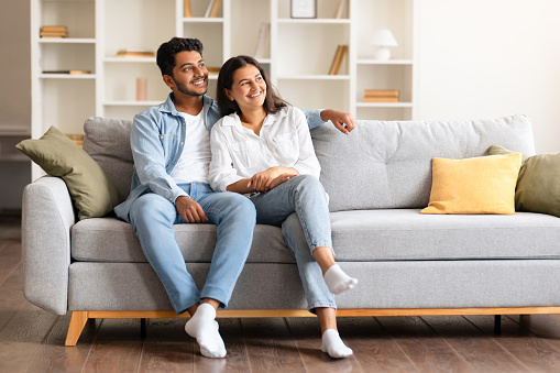 Joyful Indian couple, comfortably sitting on a sofa in their homey space, sharing a light, dreamy moment while gazing aside and sharing tender smiles, free space