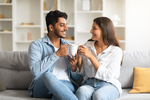Cheerful young indian couple enjoying leisurely moment on sofa, drinking coffee while having pleasant conversation, creating heartwarming, homey scenario