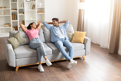 Joyful indian couple resting at home on couch with hands behind their heads, exchanging warm, loving smiles, embodying a serene and blissful domestic moment of togetherness and comfort