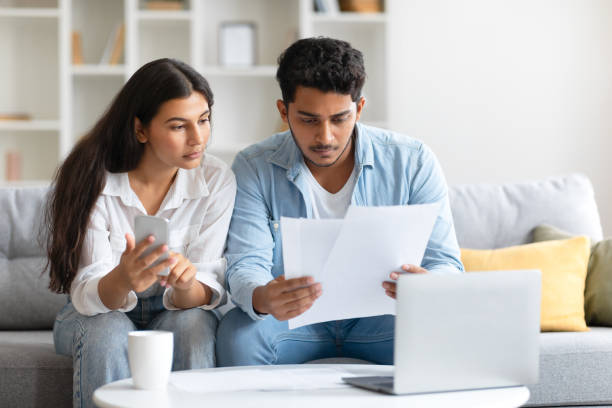 istock Indian couple focused on bills and using cellphone, sitting on sofa 1856863077