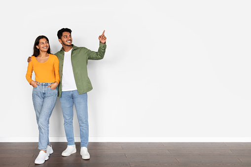 Delightful indian couple embracing and smiling while concurrently pointing to free space, expressing joy and excitement over white wall background, full length