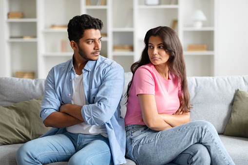 Young Indian couple sits side by side on cozy couch, their expressions contemplative, revealing moment of reflection or possible disagreement in homey setting