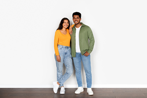 Cheerful young Indian spouses dressed in modern casual clothes, confidently stands together against clean white background, exuding warmth and camaraderie