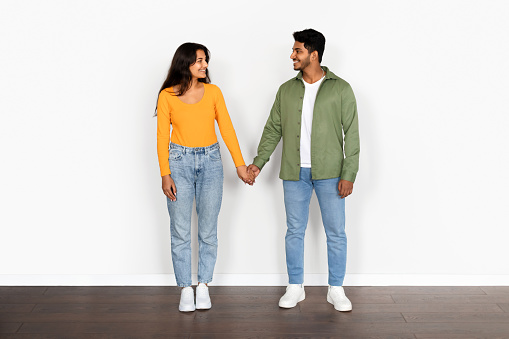 Young Indian couple, dressed in casual attire, stands against white background, holding hands and looking at each other with radiant smiles, full length
