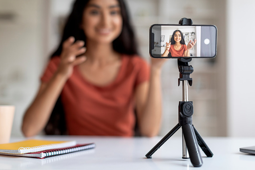 Selective focus on phone set on tripod recording millennial indian lady influencer. Young woman famous blogger shares moments using her smartphone, streaming, going live from home