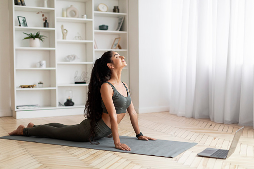 Fitness online. Athletic indian young woman doing yoga in living room, stretching on yoga mat in front of laptop, copy space. Sporty eastern lady exercising at home, using video lessons, side view