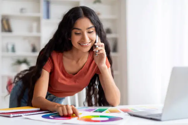 Remote Business Ideas. Young Indian Woman Freelance Designer Working With Laptop And Cellphone At Cozy Home Office, Talking With Client, Checking Color Palette, Free Space