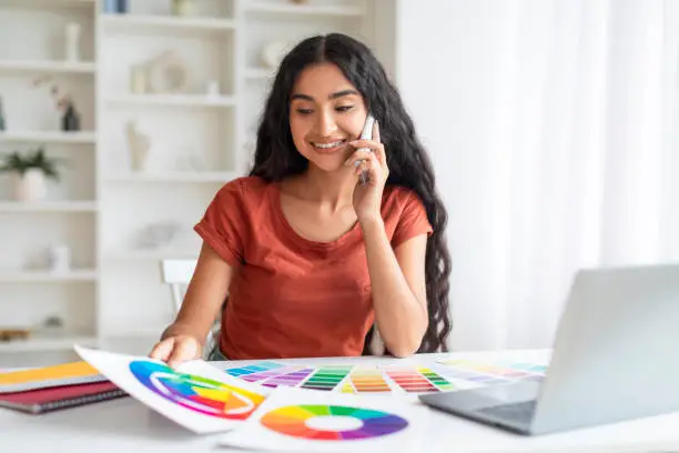 Positive creative millennial indian woman freelancer, calmly managing her design projects while balancing between laptop use and phone conversations at home office, holding color palette