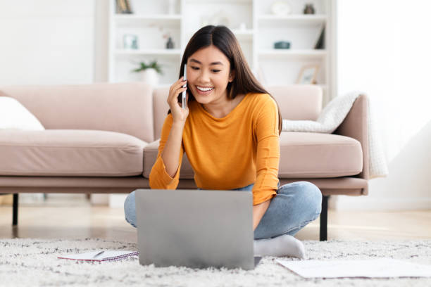 Busy young asian woman freelancer working from home Cheerful busy young asian woman freelancer working from home. Happy smiling korean lady sitting on floor in living room, have phone conversation with client, looking at laptop screen, copy space project manager remote stock pictures, royalty-free photos & images