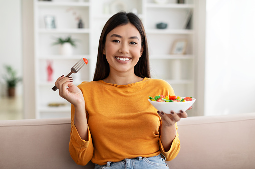 Happy attractive slim young asian woman sitting on couch at home, eating salad made of fresh organic vegetables, have healthy lunch, copy space. Healthy lifestyle, diet, nutrition concept