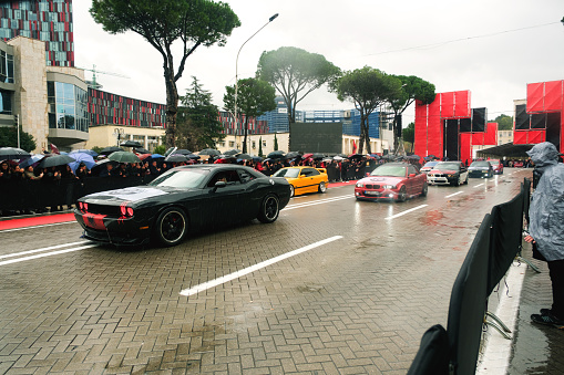 Tirana, Albania - November 28, 2023: Along Avenue of Martyrs of the Nation, a rainy Independence Day parade features a black Dodge Challenger driven by a blonde woman, backed by a lineup of BMW cars