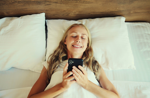 Healthy sleep, happy relaxed middle aged woman with mobile phone lying on the bed on white soft comfortable pillow