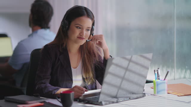Indian happy woman wearing headphone sitting on chair using laptop talking online video call indoor office. Focused female agent provide customer support service communicate with client at workplace