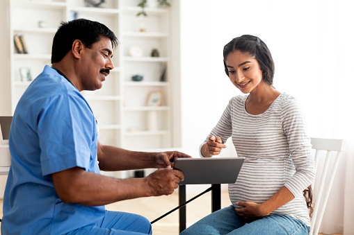 Smiling mature doctor showing digital tablet with test results to pregnant indian woman in clinic, male gynecologist in uniform consulting young expectant mother in modern hospital, giving advices