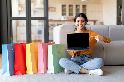 Online Shopping. Happy Indian Woman Pointing At Laptop With Blank Screen While Sitting On Floor Next To Shopper Bags At Home, Smiling Eastern Lady Recommending Website For Internet Purchases, Mockup