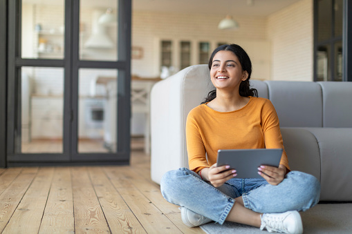 Happy Young Indian Woman With Digital Tablet Relaxing On Floor At Home, Smiling Eastern Female Using Modern Gadget And Looking Aside At Copy Space, Enjoying Technologies For Leisure