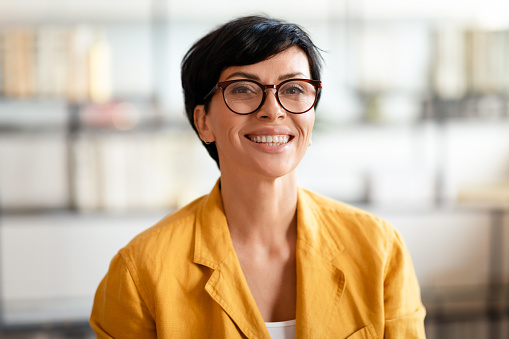 Middle aged European businesswoman in yellow jacket, smiling at camera wearing eyeglasses, sitting at office table indoor. Modern fashion and professional lifestyle