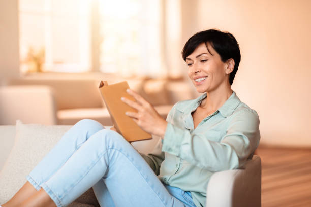istock Brunette lady smiling absorbed in reading fiction at home interior 1857116862