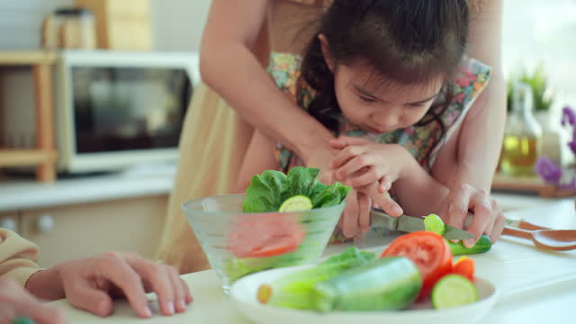 Happy Asian mother and daughter spending time together in the kitchen while they are preparing healthy salad by cutting a cucumber. Little daughter tries to cut off piece of cucumber with kitchen knife under supervision of her mom at home. Healthy food.