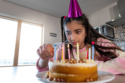 A close-up shot of an adolescent girl lighting candles on a birthday cake. She is wearing casual clothing and a pink party hat.