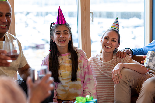 A multiracial, multi-generational family wearing casual clothing in a holiday rental accommodation located in Amble, Northumberland. They are gathered to celebrate the teenage girl's birthday and are wearing party hats in a room filled with balloons and bunting. They talk, laugh and smile with one another.